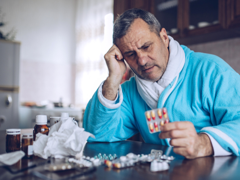 Mann mit Erkältung sitzt am Tisch und hält Tablettenpackung in der Hand